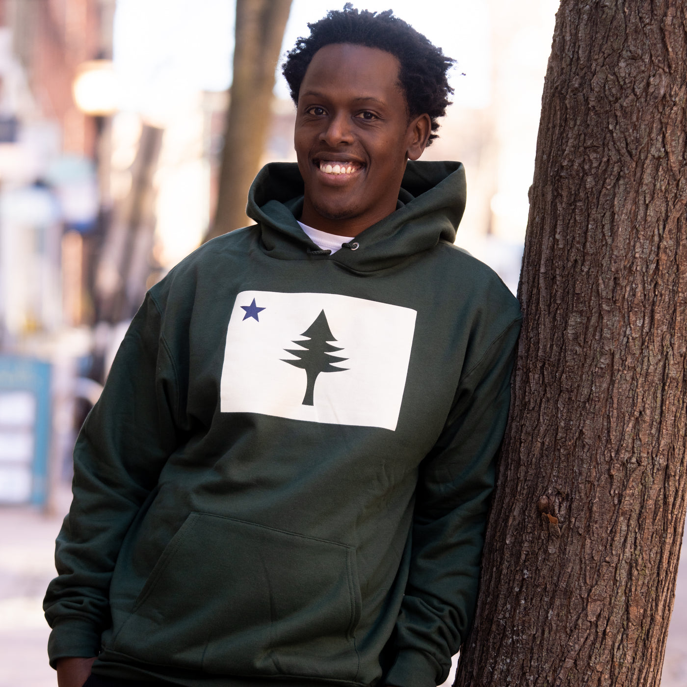 Person wearing a green hoodie with the design of the 1901 Maine state flag, leaning against a tree outdoors.