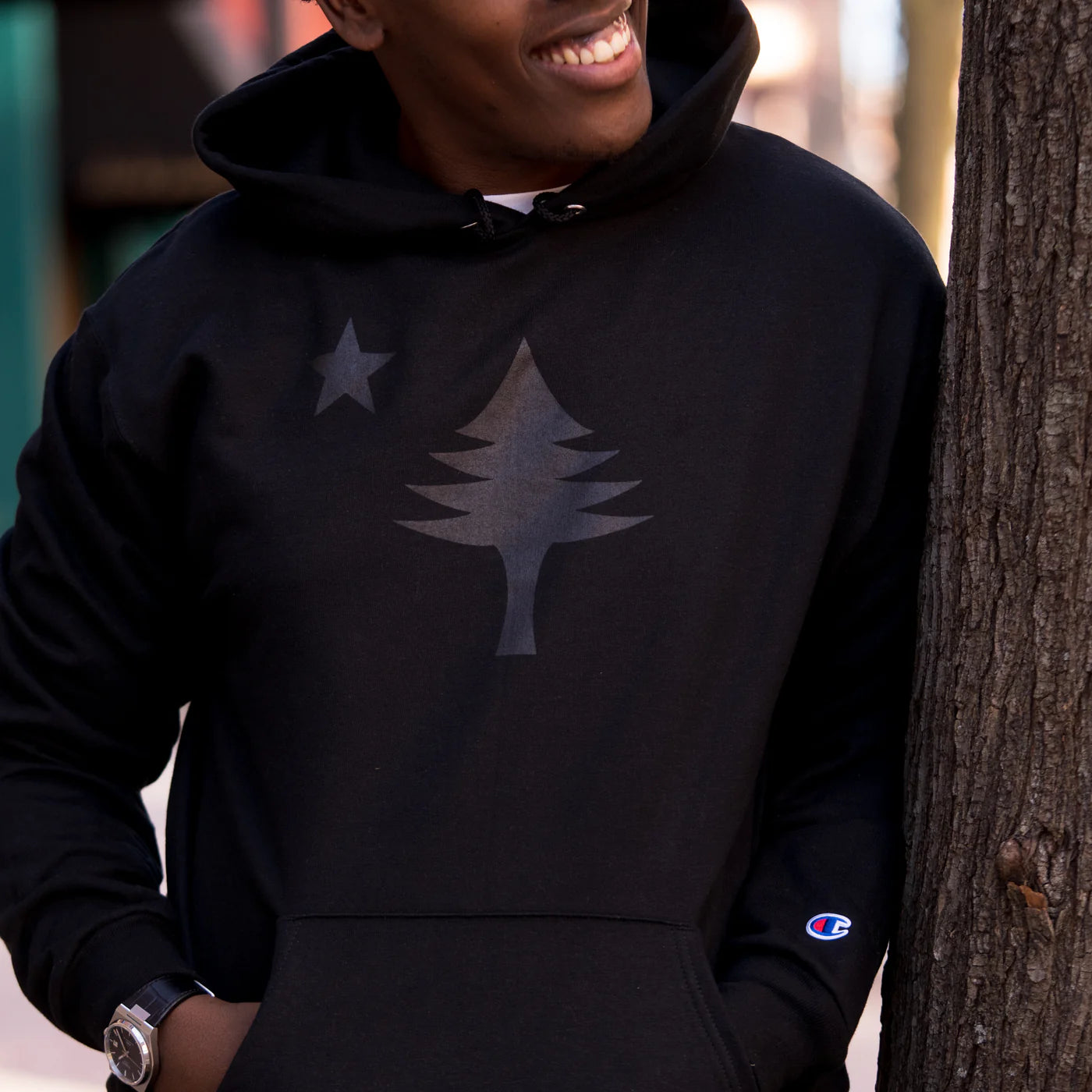 Person wearing a black hoodie with a black design of the 1901 Maine state flag, standing next to a tree.
