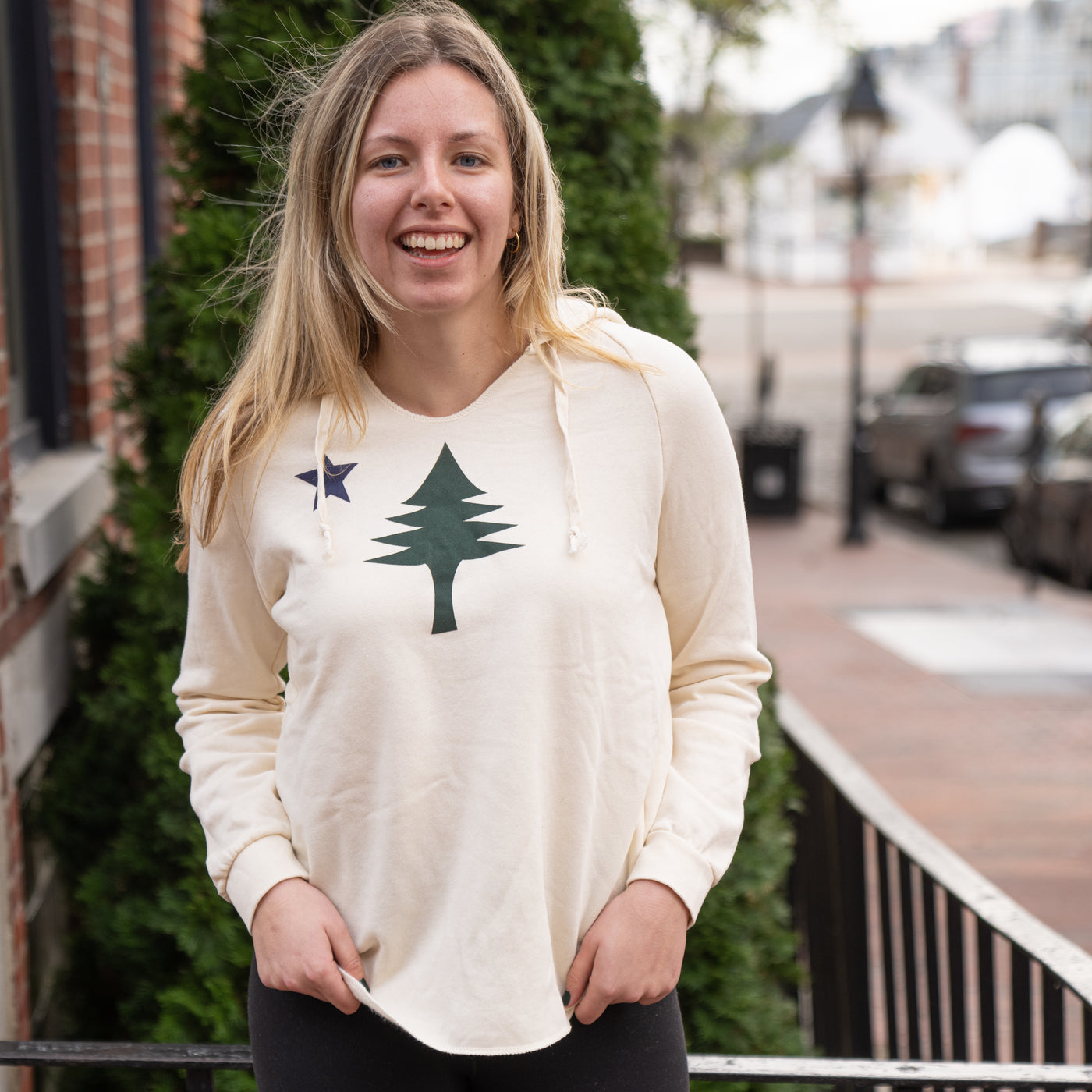 Woman wearing a white sweatshirt with the  design of the 1901 Maine state flag, standing outdoors.